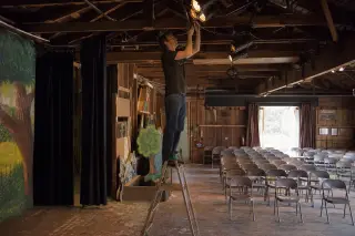 Technician adjusting stage lights in a modest village theatre, empty chairs facing the stage, natural documentary-style lighting.
