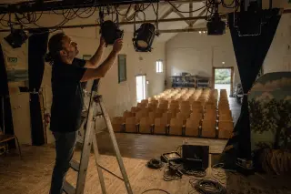 Stage technician adjusting theater lights over an empty wooden stage in a community venue, with rows of empty chairs and simple backstage equipment in view.
