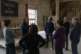 Actors rehearsing in a rustic barn with wooden beams and stone walls, standing in a circle around a simple stage set, natural daylight illuminating the scene.
