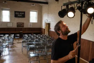 Moderate-sized community performance hall seen from the stage, empty chairs set up, technician checking stage lights, warm natural lighting, atmosphere of quiet preparation.