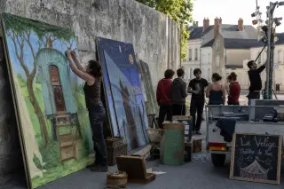 Street theatre troupe preparing scenery and props against a stone wall in a village, early morning light, performers and technician setting up for an outdoor show.
