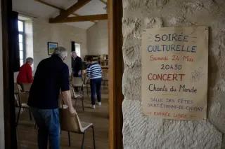 Volunteers set up chairs in a sunlit community hall with a hand-made performance poster on a stone wall.