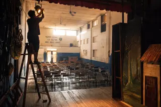Technician adjusting stage lights in a rural theater, view from stage over empty chairs with backstage elements, natural documentary lighting.