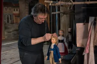 Marionnettist backstage adjusting handmade puppets under soft workspace lighting, with textured props and plain sets.