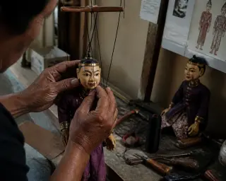 Marionnettist’s hands adjusting wooden puppets backstage with tools on a workbench, lit by natural light in a modest theater environment.