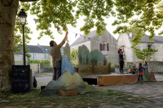 An artist arranges wooden set pieces on a small outdoor stage in a village square, early morning light, children and volunteers helping, stone houses and plane trees in the background.
