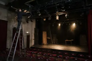 Lighting technician adjusting stage spotlights on a ladder in a modest local theater, empty chairs below, soft rehearsal lighting, backstage atmosphere.