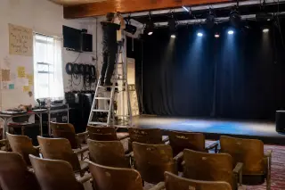 Technician on ladder adjusting stage lighting in an empty community theater, wooden chairs visible, soft morning light and stage lamps, backstage atmosphere with technical gear