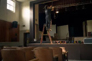 Technician adjusts stage lights in a small community theater with empty seats, morning light streaming in, evoking backstage activity before a show.