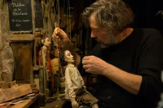 Puppeteer backstage preparing handcrafted puppets in soft rehearsal lighting, close-up on hands at work, intimate rural theater with artisanal atmosphere.