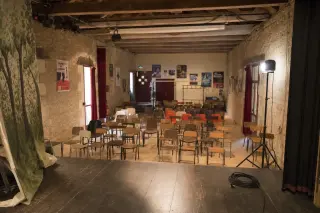Empty communal theater in Indre-et-Loire seen from stage, rows of simple chairs awaiting audience, warm rehearsal lighting, visible technical equipment and stone walls with local posters.
