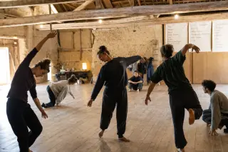 Rehearsal of a small contemporary dance company in a rustic, repurposed barn, dancers practicing under wooden beams with natural and soft rehearsal lighting.