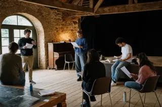 Actors rehearsing in a rustic barn converted into a small cultural space, with visible wooden beams and stone walls, under soft natural and artificial light